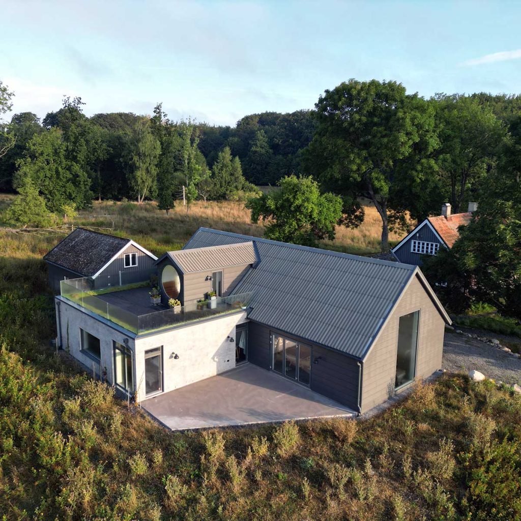 An aerial photo of Marie Louise Kold's studio, a large dark grey building with a tall gable window and a lighter grey annex with a roof terrace and a large round window, a forest in the background.