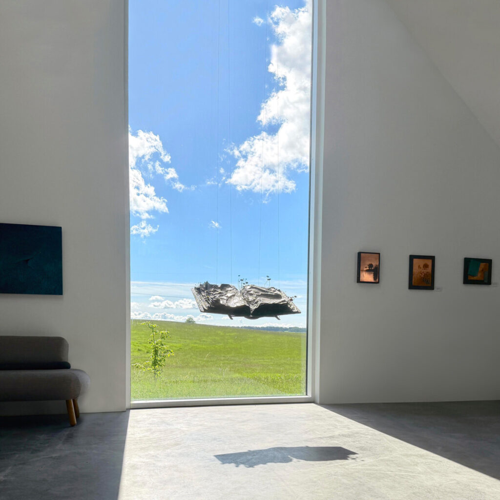 The Book of Alchemical Secrets hanging in the studio, suspended in front of a tall window. The studio is a bright white space with a high ceiling and the view through the window shows a green field and blue sky