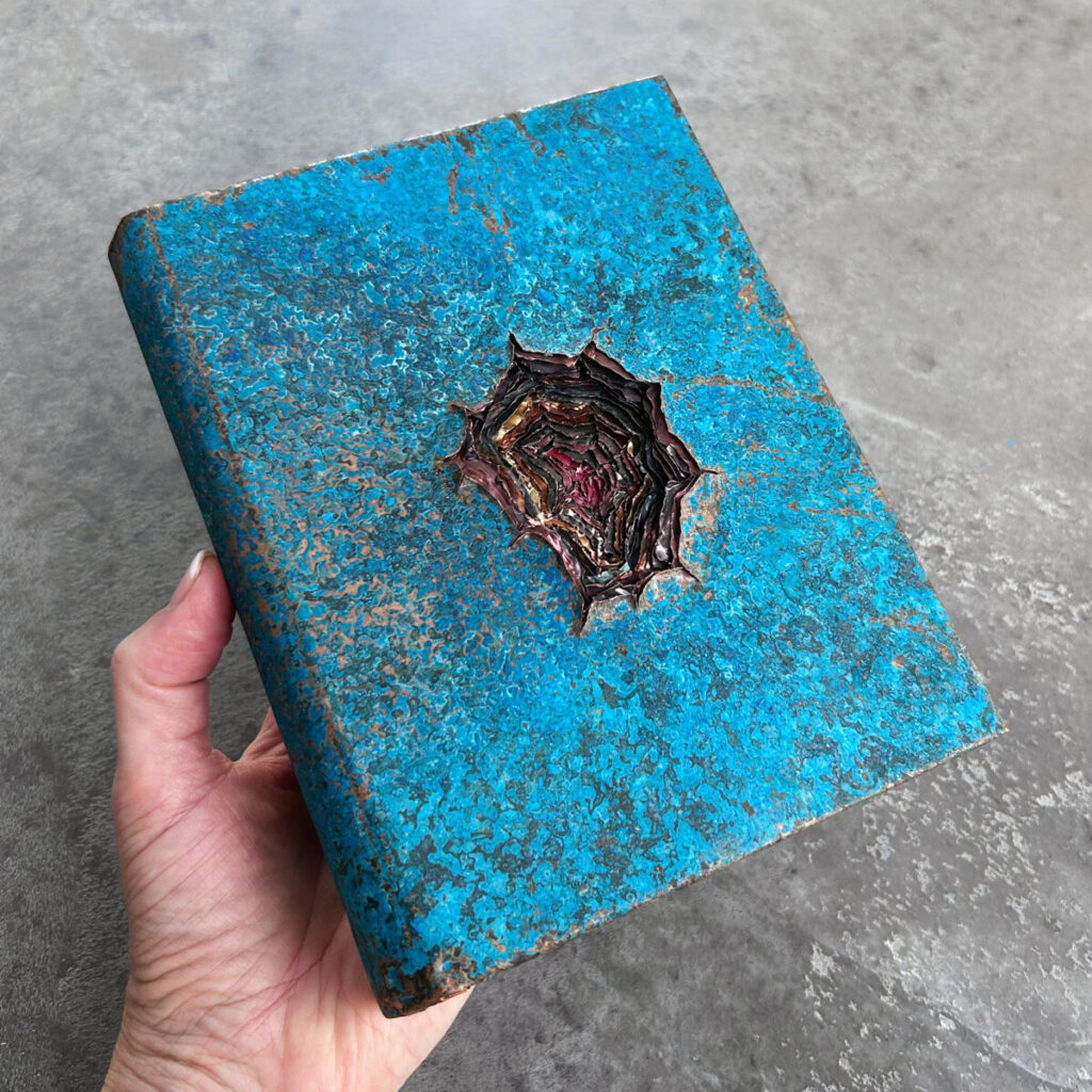 Dangerous Knowledge held in hand, a turquoise patinated copper book with a deep wound in the cover