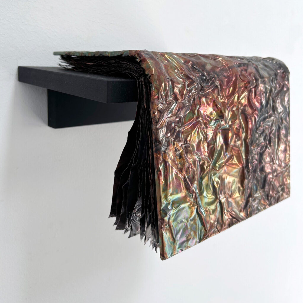 Guide to the Wilderness, a patinated copper artist's book draped over a black wall shelf, patinated copper pages cascading over the edge