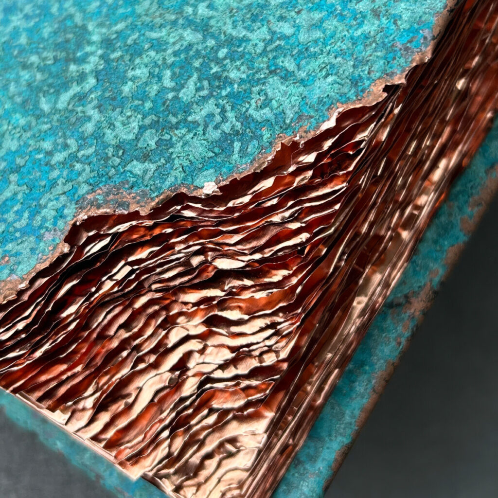 Detail of the torn corner of Wilderness Survival Manual, the rippled bare copper pages set against the turquoise cover